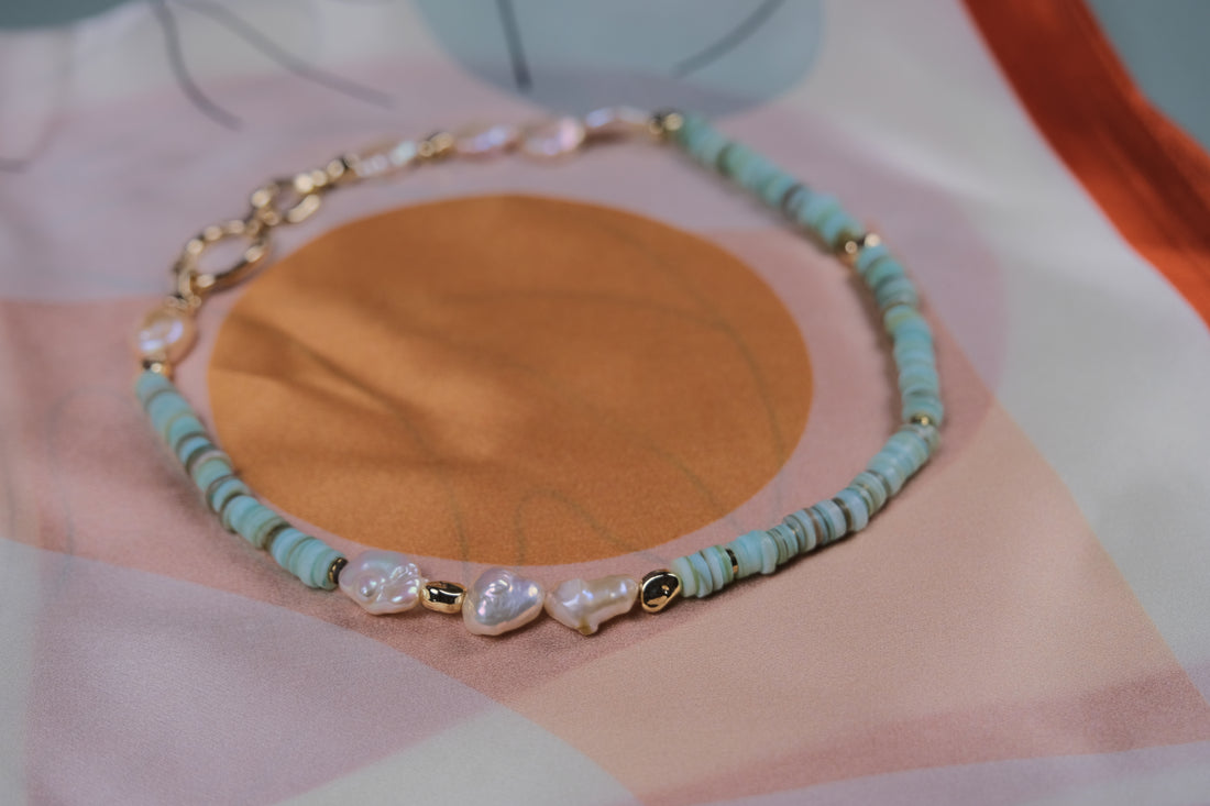 Un collier composé de perles baroques, de turquoise naturelle, et d’or laminé 14 carats, délicatement posé sur une écharpe en soie. L’association des perles et de la turquoise évoque une élégance joyeuse, tandis que le contraste entre les pierres et la texture douce de la soie met en valeur la beauté naturelle du collier.
